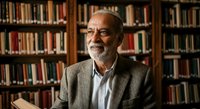 Senior man with gray beard and glasses wearing olive cardigan over light blue shirt, seated in library with wooden bookshelves background, suitable for education, academia, and professional portraits.