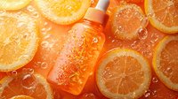 Orange cosmetic serum bottle with fresh citrus slices and water drops on coral background, skincare product photography for beauty and wellness marketing.