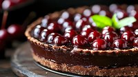 Dark chocolate tart topped with fresh cherries and mint leaves on rustic wooden surface, rich ganache filling creates luxurious dessert presentation.