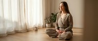 Young Caucasian woman in beige linen outfit practicing mindful meditation sitting cross-legged on wooden floor in bright natural light room with plants.