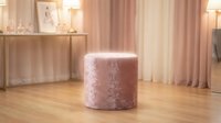 Soft pink velvet cylindrical ottoman pouf in modern living room with wooden floor, curtains, and decorative shelving for interior design projects.