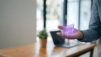 Young professional man holding glowing purple holographic pyramid above tablet device at wooden desk, representing digital innovation, technology advancement, and futuristic business solutions.