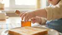 Young child with light skin tone spreading honey or jam on bread with a knife during breakfast or snack preparation at home.