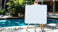 Blank white canvas on wooden easel by swimming pool in tropical resort setting for art classes, workshops, and creative outdoor painting sessions.
