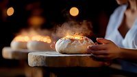 Fresh baked bread loaf with steam rising on wooden cutting board in dark moody setting, female hands holding artisanal sourdough in rustic kitchen atmosphere.