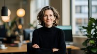 Confident mature businesswoman with curly hair in black turtleneck standing with arms crossed in modern office environment with natural lighting.