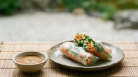 Fresh rice paper rolls filled with colorful vegetables served on a ceramic plate with peanut dipping sauce on a bamboo mat.