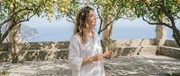 Woman in white linen shirt standing under olive trees in Mediterranean landscape, holding wine glass, relaxed summer lifestyle setting.