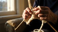 Elderly hands knitting with wooden needles and thick cream wool yarn by a sunlit window, capturing cozy handcraft and traditional hobby.