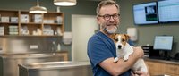 Middle-aged Caucasian male veterinarian with glasses and beard holding small Jack Russell terrier dog in modern veterinary clinic examination room.