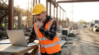 Construction worker in safety vest and hard hat using laptop at building site during golden hour sunset for project management and planning.