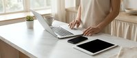 Woman working on laptop at white desk with tablet and plant in bright modern home office workspace for remote work productivity and business tasks.