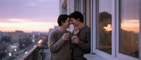 Young couple embracing on urban balcony at sunset, sharing intimate moment with cityscape and golden hour lighting in background, romantic relationship concept.