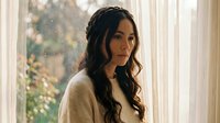 Young Hispanic woman with braided hair stands by a sunlit window, wearing a cozy cream sweater, with a thoughtful, melancholic expression.