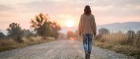 Woman in casual clothing walking alone on rural dirt road at sunset with mountains in distance, representing solitude, journey, and peaceful outdoor lifestyle.