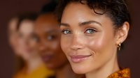 Young mixed race woman with natural curly hair and glowing skin smiling warmly at camera, wearing orange top. Soft beauty portrait with blurred background.
