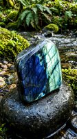 Polished labradorite gemstone displaying blue and green iridescence sits on moss-covered rock beside forest stream with lush ferns creating mystical atmosphere.