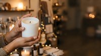 Hands holding lit white scented candle with warm bokeh lights in cozy home interior, perfect for wellness, relaxation, and holiday decor concepts.