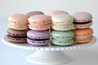 Colorful French macarons in pastel shades of pink, purple, mint green, and peach arranged on white ceramic cake stand, showcasing delicate meringue cookies.