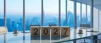 Modern office interior with wooden blocks displaying 2027 on glass desk overlooking city skyline through floor-to-ceiling windows at dusk.