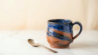 Handmade ceramic coffee mug with blue and orange glazed finish on white marble surface with copper spoon for morning beverage and kitchen decor.