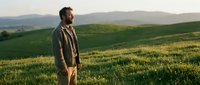 Middle-aged Caucasian male farmer or agricultural professional standing in expansive green rolling hills and meadow landscape during golden hour, wearing casual work clothing and jacket, surveying farmland or countryside terrain.