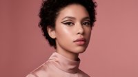 Young mixed-race woman with curly hair and winged eyeliner wearing pink turtleneck against coral background for beauty and fashion concepts.