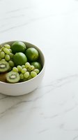 Fresh green fruits including limes, kiwis and grapes arranged in white bowl on marble surface. Healthy eating and nutrition concept for food styling and culinary design projects.