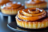 Caramel glazed mini tart with swirled topping on dark plate, close-up view showing glossy surface and flaky pastry crust, for food and dessert marketing.
