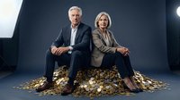 Professional senior business couple sitting on pile of golden coins representing wealth and financial success in corporate environment.