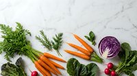 Fresh organic vegetables including carrots, red cabbage, radishes and leafy greens arranged on white marble surface for healthy cooking and nutrition.