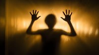 Silhouette of person with hands pressed against frosted glass in warm golden light creating dramatic shadow effect for horror or mystery concepts.