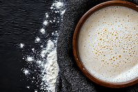 Creamy white milk beverage with golden bubbles in brown ceramic cup on dark background with scattered flour, top view composition for food blogs.