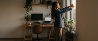 Young woman with long dark hair and medium skin tone stretching at her home office desk, reaching toward window with arm extended, wearing casual blazer, taking break from work.