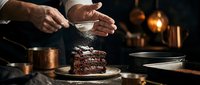 Pastry chef dusting powdered sugar onto chocolate cake slice in warm kitchen lighting, baking and dessert preparation.
