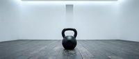 Black kettlebell weight on dark wooden floor in minimalist gym studio with white walls and ceiling light. Fitness training equipment.