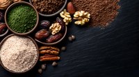 Superfood ingredients in wooden bowls including green powder, cocoa, nuts, dates and seeds arranged on dark slate background for healthy cooking and nutrition.