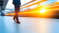 Motion blur of passing train with silhouette of person walking on platform creates dynamic composition showing speed, movement and urban transit atmosphere.