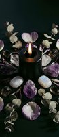 Black candle burning surrounded by amethyst crystals, moonstone gems and eucalyptus leaves on dark background for spiritual meditation and healing rituals.