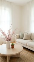 Minimalist living room interior with beige sofa, round wooden coffee table, cherry blossom branches in vase, natural light through white curtains.