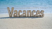 Wooden letters spelling Vacances on sandy beach with turquoise ocean waves, market.