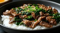 Savory beef strips with fresh herbs and sesame seeds served over steamed white rice in dark bowl, perfect for Asian cuisine menu or cooking blog content.