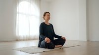 Young Asian woman in black athletic wear practicing meditation in lotus position on yoga mat in bright minimalist room with natural window light.