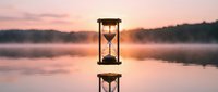 Hourglass reflecting on calm water surface during golden sunset with misty lake and forest silhouette creating peaceful time concept for meditation and mindfulness.