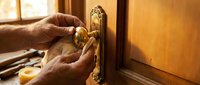 Professional locksmith hands installing brass door handle on wooden door with vintage ornate backplate in warm interior lighting for home improvement projects.