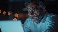 Senior businessman working late at laptop in dark office with blue light illumination and bokeh background, focused expression showing dedication.