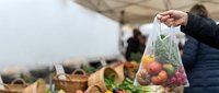 Farmer holding mesh bag of fresh tomatoes at outdoor market or farm stand with harvest baskets in background.
