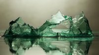 Shattered green glass fragments creating mountain-like shapes with reflection on smooth surface against blurred background, dramatic lighting creates ethereal atmosphere.