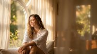 Young woman with curly hair in white shirt sitting by window with warm sunlight streaming through, creating peaceful morning atmosphere for lifestyle content.