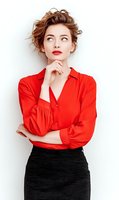 Young woman with auburn hair in red blouse and black skirt looking up thoughtfully with hand on chin against white background. Perfect for fashion or business concepts.